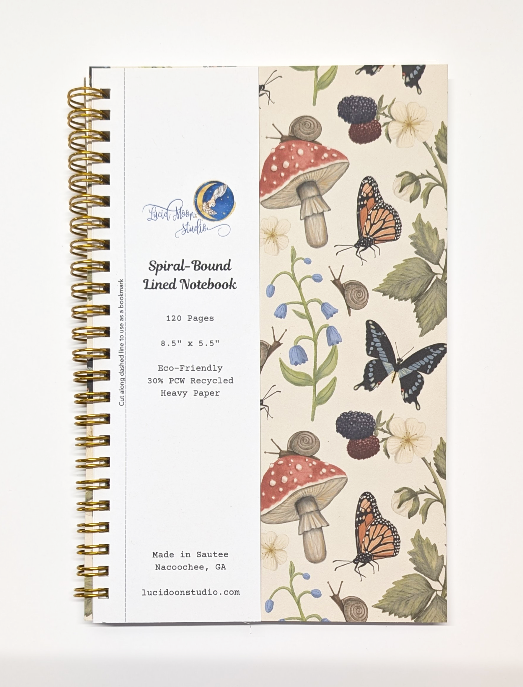 Gold spiral notebook with nature-themed cover design featuring mushrooms, butterflies, snails, frogs, blackberry branches, and bluebell flowers on a beige background with a detachable label.