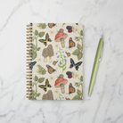 Gold spiral notebook with nature-themed cover design featuring mushrooms, butterflies, snails, frogs, blackberry branches, and bluebell flowers on a beige background.