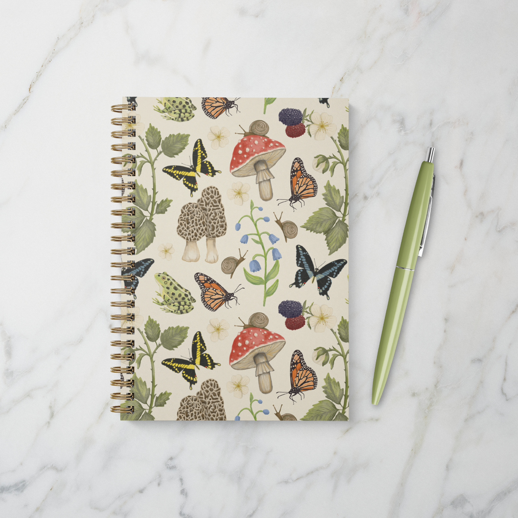 Gold spiral notebook with nature-themed cover design featuring mushrooms, butterflies, snails, frogs, blackberry branches, and bluebell flowers on a beige background.