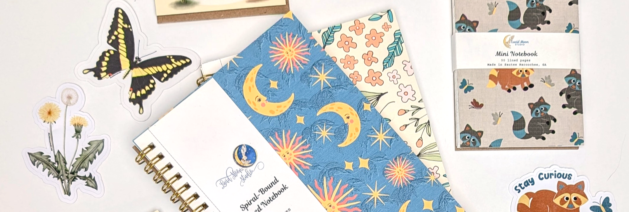 An assortment of products including two spiral notebook with celestial and floral patterns, a mini spiral notebook with a raccoon pattern, and three vinyl stickers with a swallowtail butterfly, dandelions, and a raccoon illustration that says stay curious.