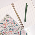 Notebook with floral cover, spiral binding, and green pen on a white surface