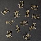 A set of gold-colored metal paperclips shaped like various cartoon animals and objects, including a cat, bow, cherries, fountain pen, dachshund, and envelope, arranged on a dark textured background.