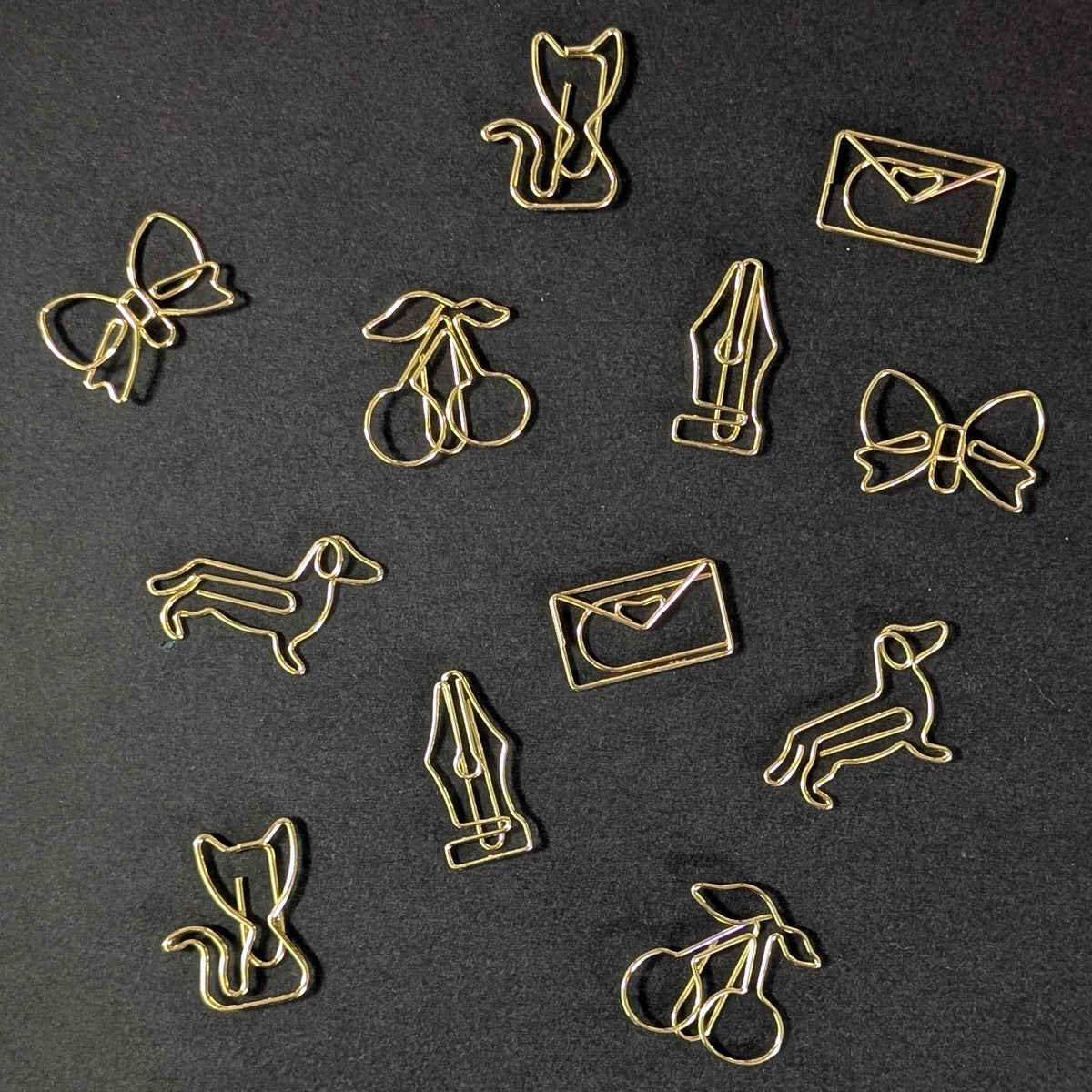 A set of gold-colored metal paperclips shaped like various cartoon animals and objects, including a cat, bow, cherries, fountain pen, dachshund, and envelope, arranged on a dark textured background.