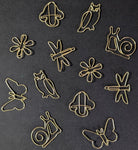A set of small gold metal wire paper clips shaped like owls, butterflies, snails, flowers, and a clover, arranged on a dark textured background.