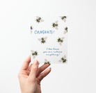 Hand holding a Greeting card with hand-drawn bee illustrations on a white background with blue text that says "Congrats! I bee-lieve you can achieve anything!" 