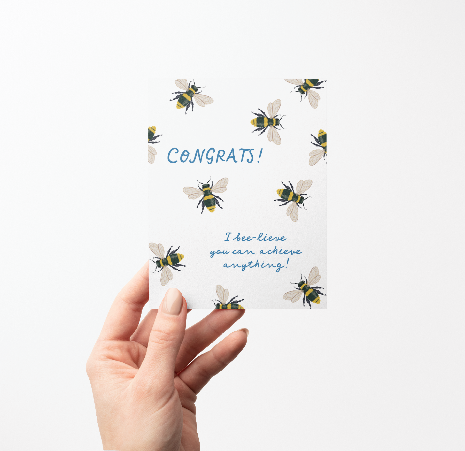Hand holding a Greeting card with hand-drawn bee illustrations on a white background with blue text that says "Congrats! I bee-lieve you can achieve anything!" 