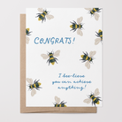 Greeting card with hand-drawn bee illustrations on a white background with blue text that says "Congrats! I bee-lieve you can achieve anything!" with a brown kraft envelope.