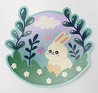 A hand-drawn matte die cut vinyl sticker featuring a cute Kawaii bunny sitting in a meadow surrounded by leaves, flowers, a pink butterfly, a heart, and a cloud in the background.
