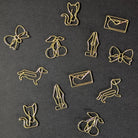 A set of gold-colored metal paperclip charms shaped like a cat, bow, cherries, fountain pen, dachshund, rabbit, and envelope, arranged on a dark textured background.