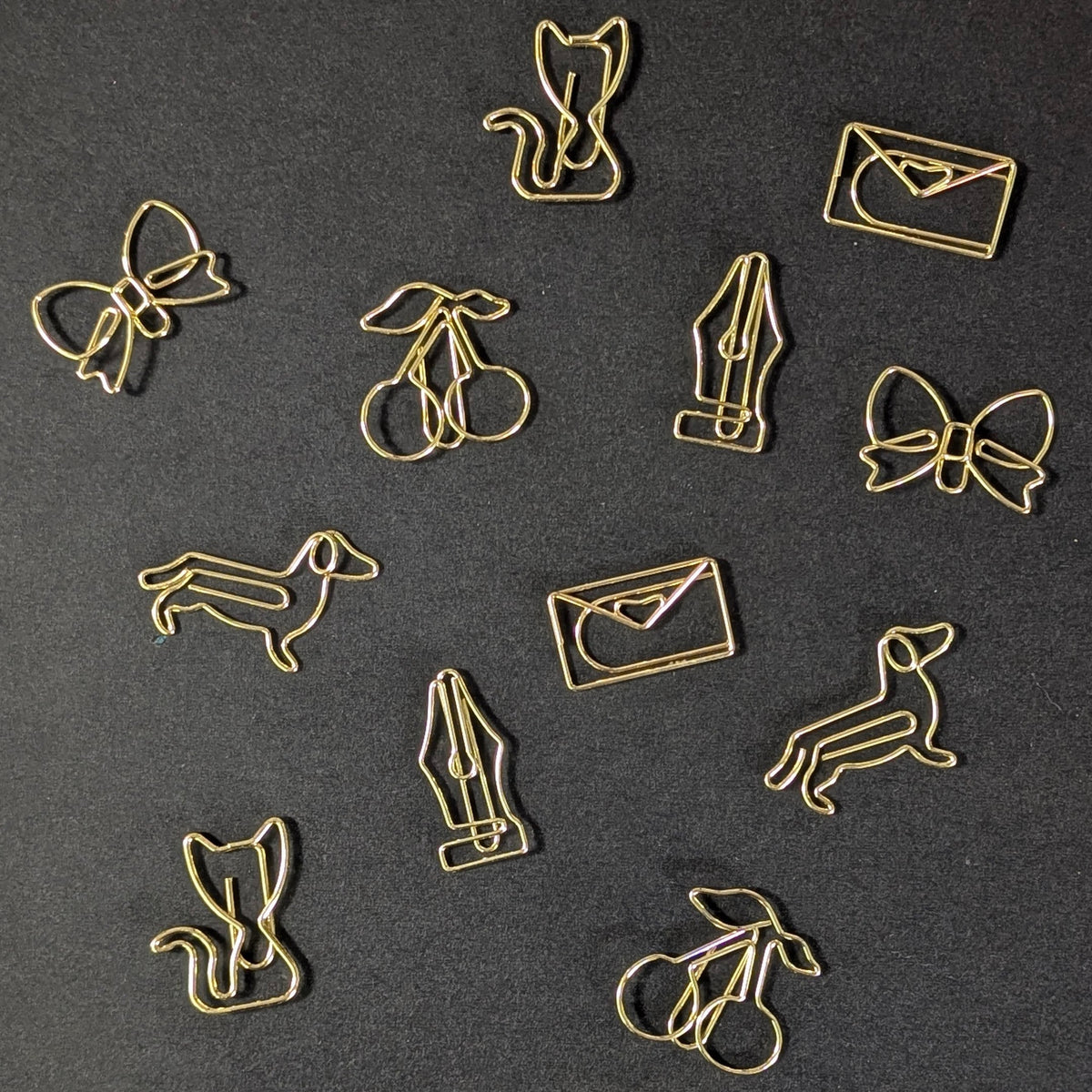 A set of gold-colored metal paperclip charms shaped like a cat, bow, cherries, fountain pen, dachshund, rabbit, and envelope, arranged on a dark textured background.