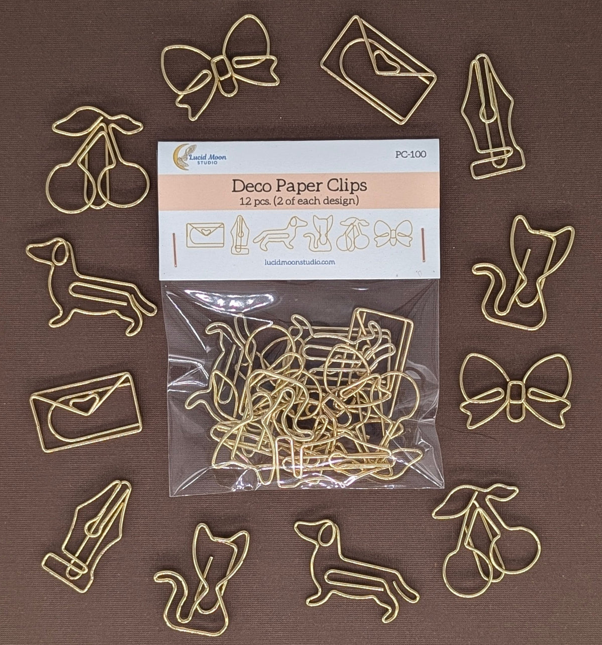 A set of 12 gold-colored metal wire paper clips shaped like cherries, bows, envelopes, a fountain pen, a dog, a squirrel, and a cherry, arranged around a clear plastic bag of the product on a solid brown background.