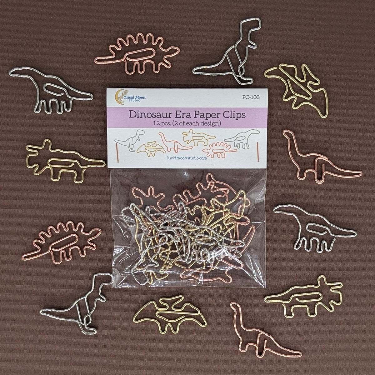 A pack of dinosaur-shaped paper clips arranged around their retail packaging on a solid brown background. The paper clips are made of thin metal wire in various colors including gold, silver, pink, and green, while the packaging is labeled 'Dinosaur Era Paper Clips'.