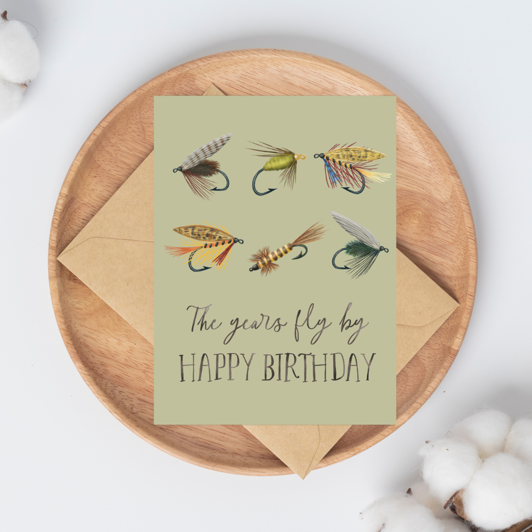 Birthday card with 6 fishing fly illustrations and text "The years fly by HAPPY BIRTHDAY" on a light green background on top of a brown kraft envelope.