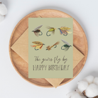 Birthday card with 6 fishing fly illustrations and text "The years fly by HAPPY BIRTHDAY" on a light green background on top of a brown kraft envelope.