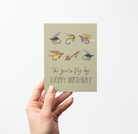 Hand holding a Birthday card with 6 fishing fly illustrations and text "The years fly by HAPPY BIRTHDAY" on a light green background.