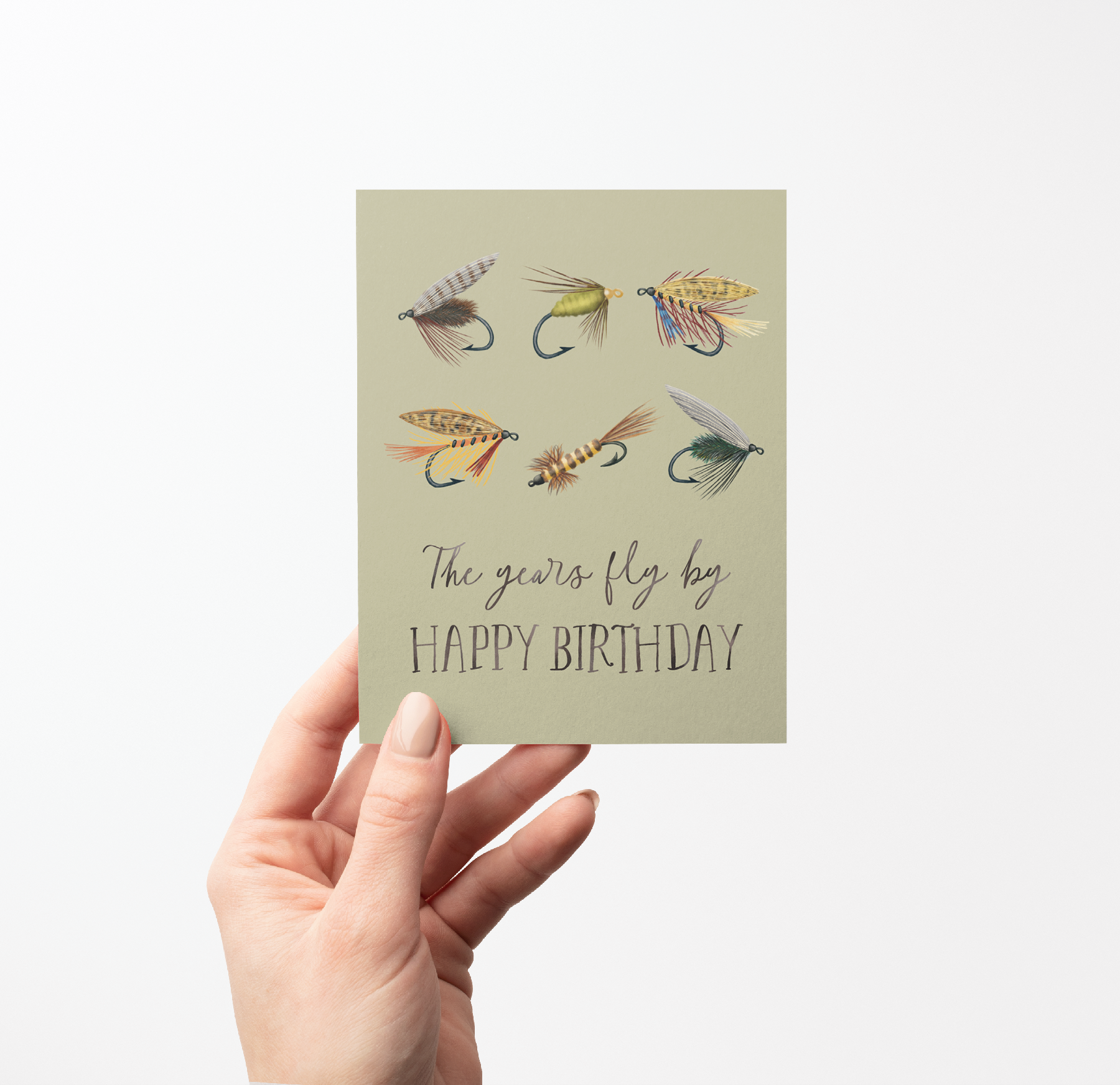Hand holding a Birthday card with 6 fishing fly illustrations and text "The years fly by HAPPY BIRTHDAY" on a light green background.