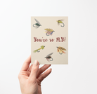 Hand holding a love/friendship card with 6 fishing fly illustrations and text "You're so FLY!" on a tan background.