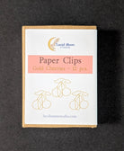 Gold Cherries paper clip set in a kraft paper box with a white label, photographed on a dark textured background.