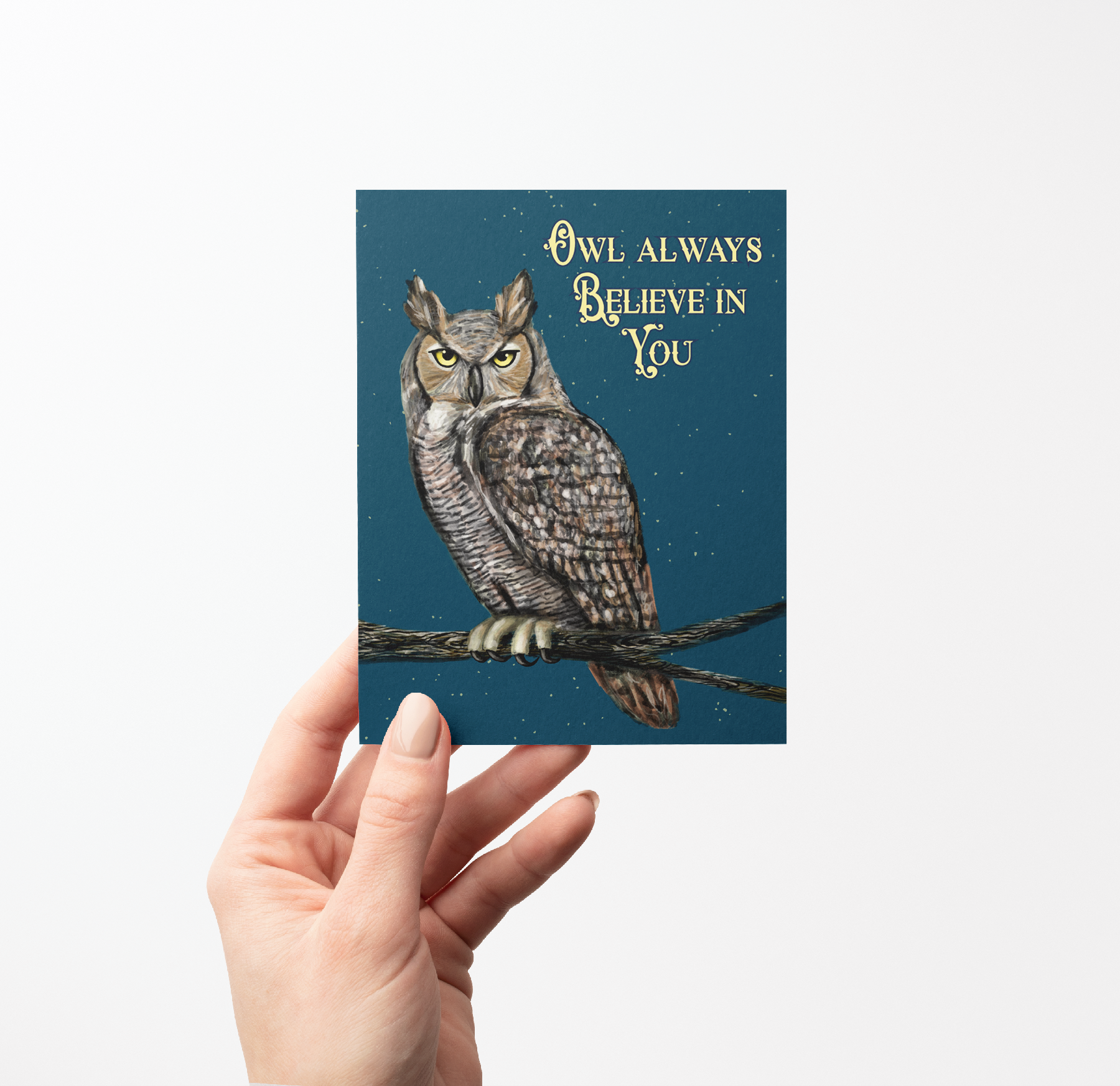 Hand holding greeting card with a painted Great Horned owl on a branch illustration on a dark blue background with stars.
