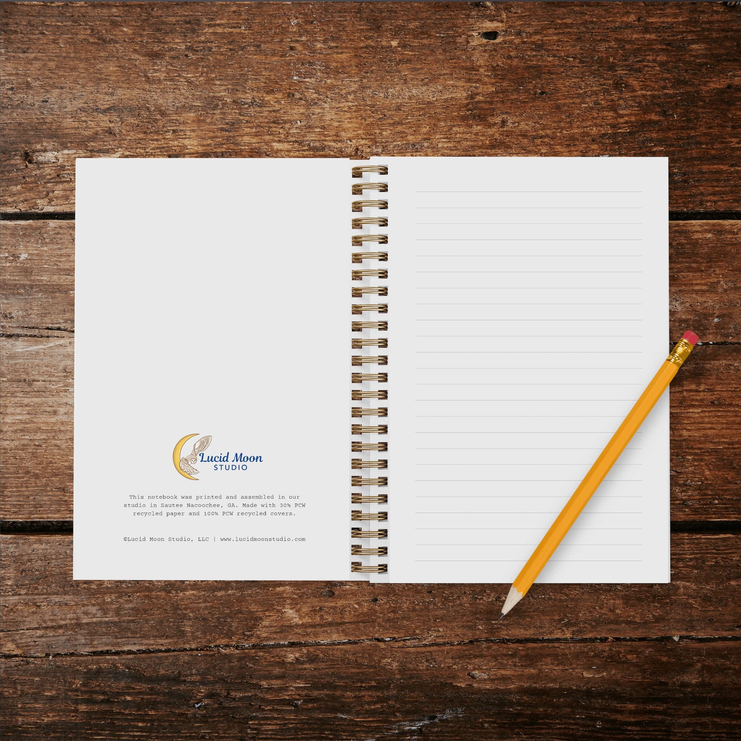 A photo of an opened wire spiral-bound notebook showing inside cover with Lucid Moon Studio logo and info about the notebook and lined pages on a wood background with a pencil.