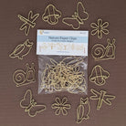 A set of 12 gold-colored metal paper clips shaped like nature items (snails, butterflies, owls, flowers, dragonflies, acorns) arranged around a clear plastic bag of the product on a solid brown background.