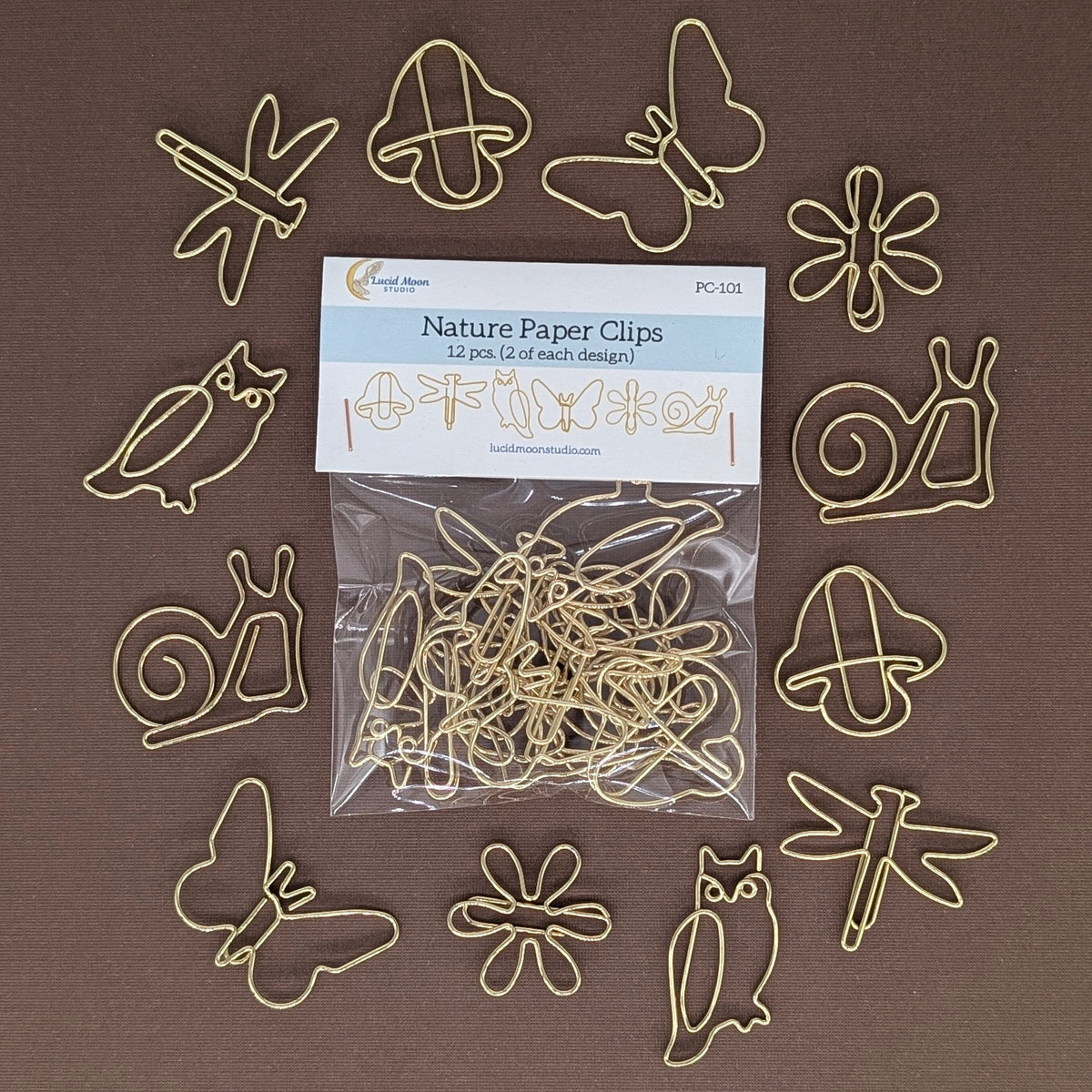 A set of 12 gold-colored metal paper clips shaped like nature items (snails, butterflies, owls, flowers, dragonflies, acorns) arranged around a clear plastic bag of the product on a solid brown background.