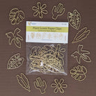 A 12-pack of gold metal paper clips shaped like plants, flowers, and leaves, shown in its clear plastic packaging with a white header card, surrounded by the unclipped clips on a solid brown background.