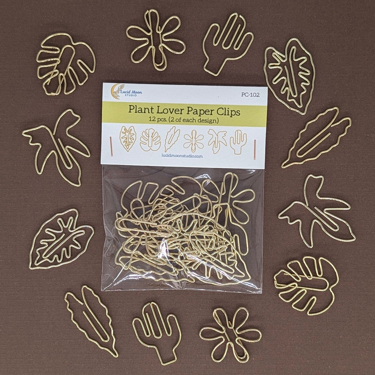 A 12-pack of gold metal paper clips shaped like plants, flowers, and leaves, shown in its clear plastic packaging with a white header card, surrounded by the unclipped clips on a solid brown background.