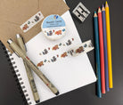 A roll of washi tape in a paper wrapper and 2 strips of washi tape adorned with hand drawn brown and gray raccoons and colorful butterflies on a white background on a notebook and kraft envelope along with pens, colored pencils, and a pencil sharpener.