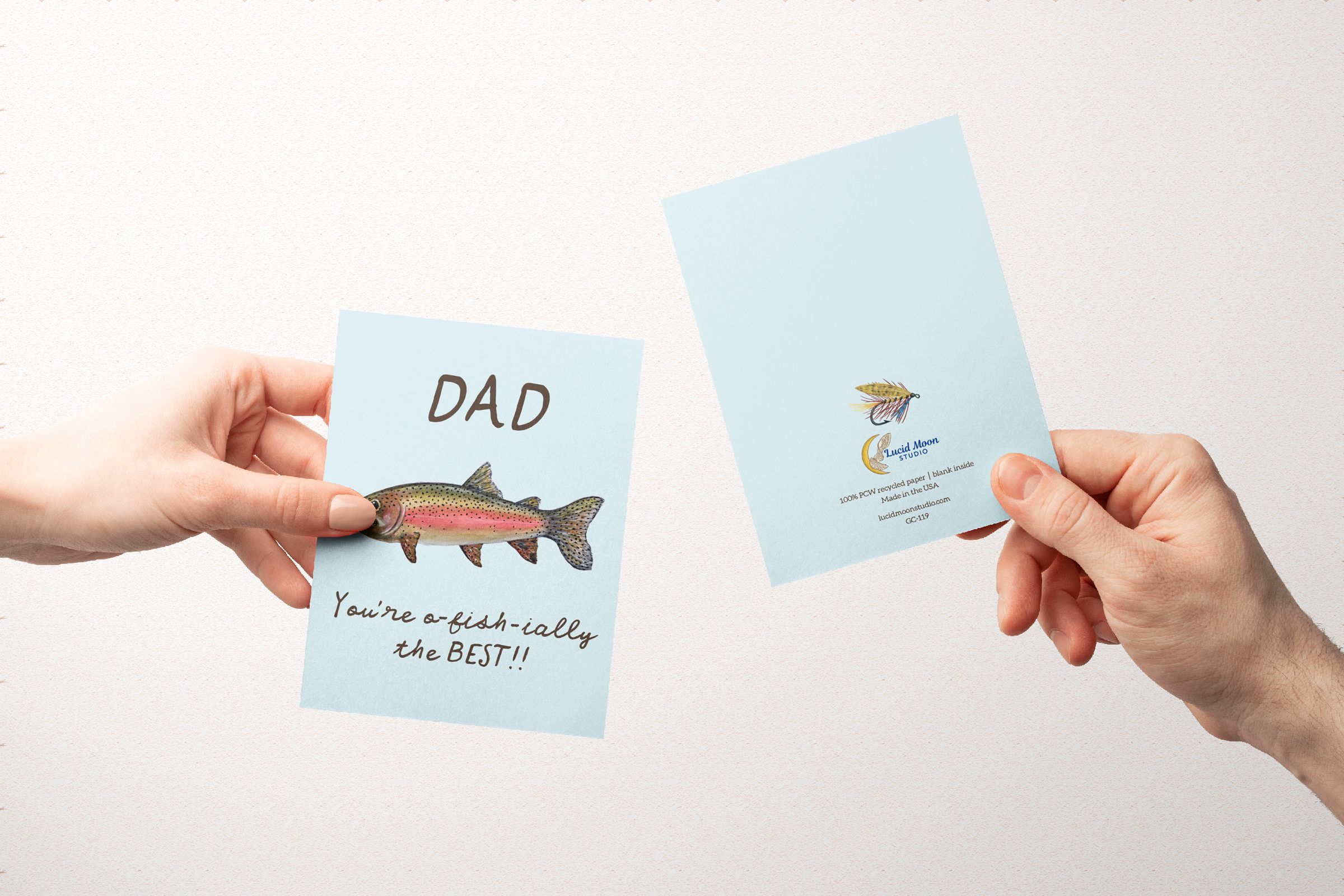 Two hands holding greeting cards with the front and back of a Greeting card with a watercolor rainbow trout illustration and the text "Dad You're o-fish-ially the BEST" on a light blue background.