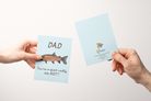 Two hands holding greeting cards with the front and back of a Greeting card with a watercolor rainbow trout illustration and the text "Dad You're o-fish-ially the BEST" on a light blue background.