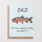 Greeting card with a watercolor rainbow trout illustration and the text "Dad You're o-fish-ially the BEST" on a light blue background on top of a brown kraft envelope.