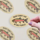 Oval-shaped sticker with a watercolor rainbow trout illustration and the words "trout don't live in ugly places" on a tan background.