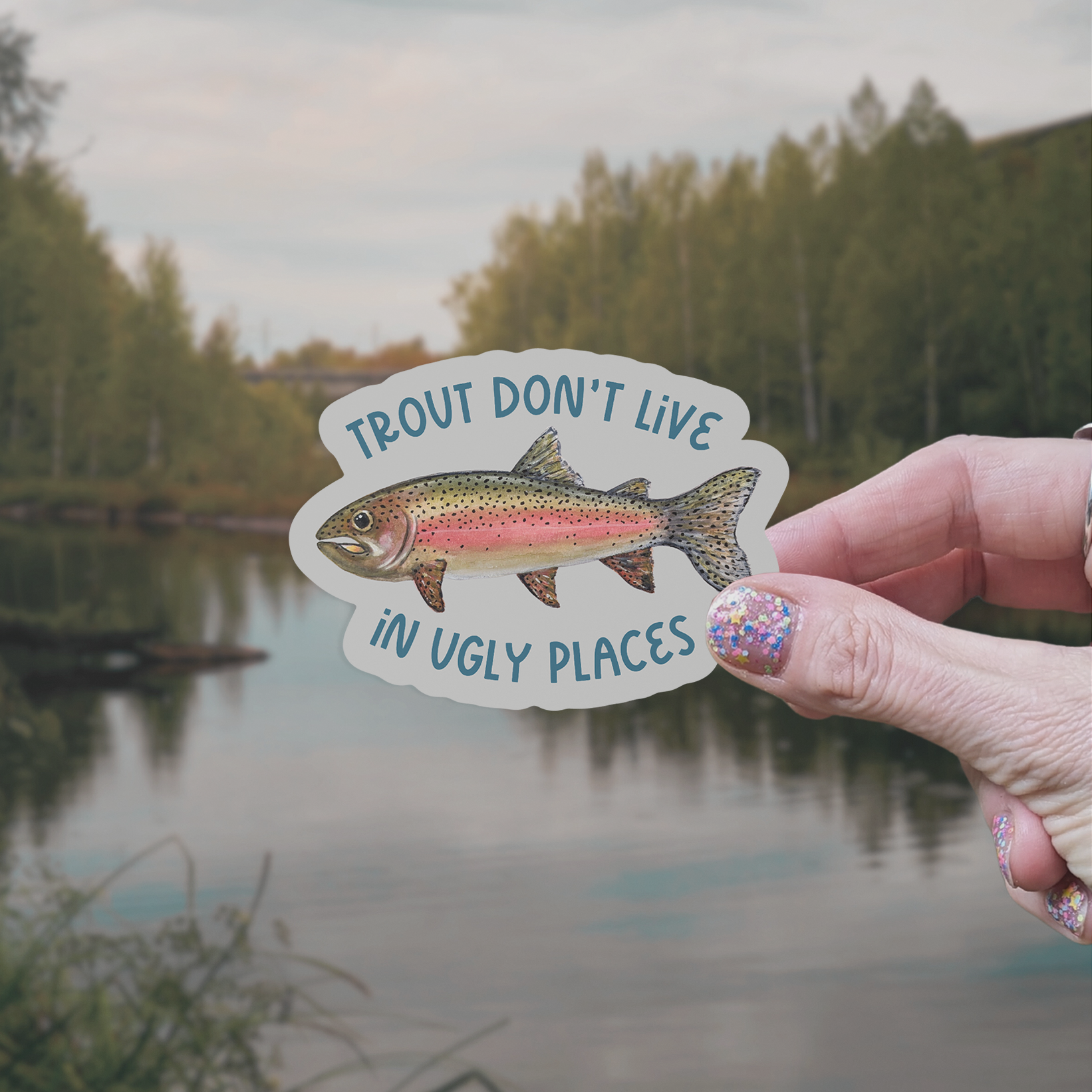 Hand holding a Die cut sticker with a watercolor rainbow trout illustration and the text "Trout don't live in ugly places" on a grey background