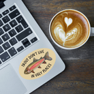 Laptop with an oval-shaped sticker with a watercolor rainbow trout illustration and the words "trout don't live in ugly places" on a tan background stuck on it next to a cup of coffee with latte art on a wooden surface.