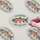 Oval-shaped sticker with a watercolor rainbow trout illustration and green text "Fish Around and Find Trout" on a grey background