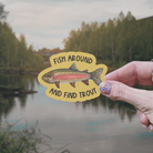 Hand holding a die cut vinyl sticker with a watercolor rainbow trout illustration on a mustard-colored background with the words "Fish around and find trout" in front of a lake and trees in the background
