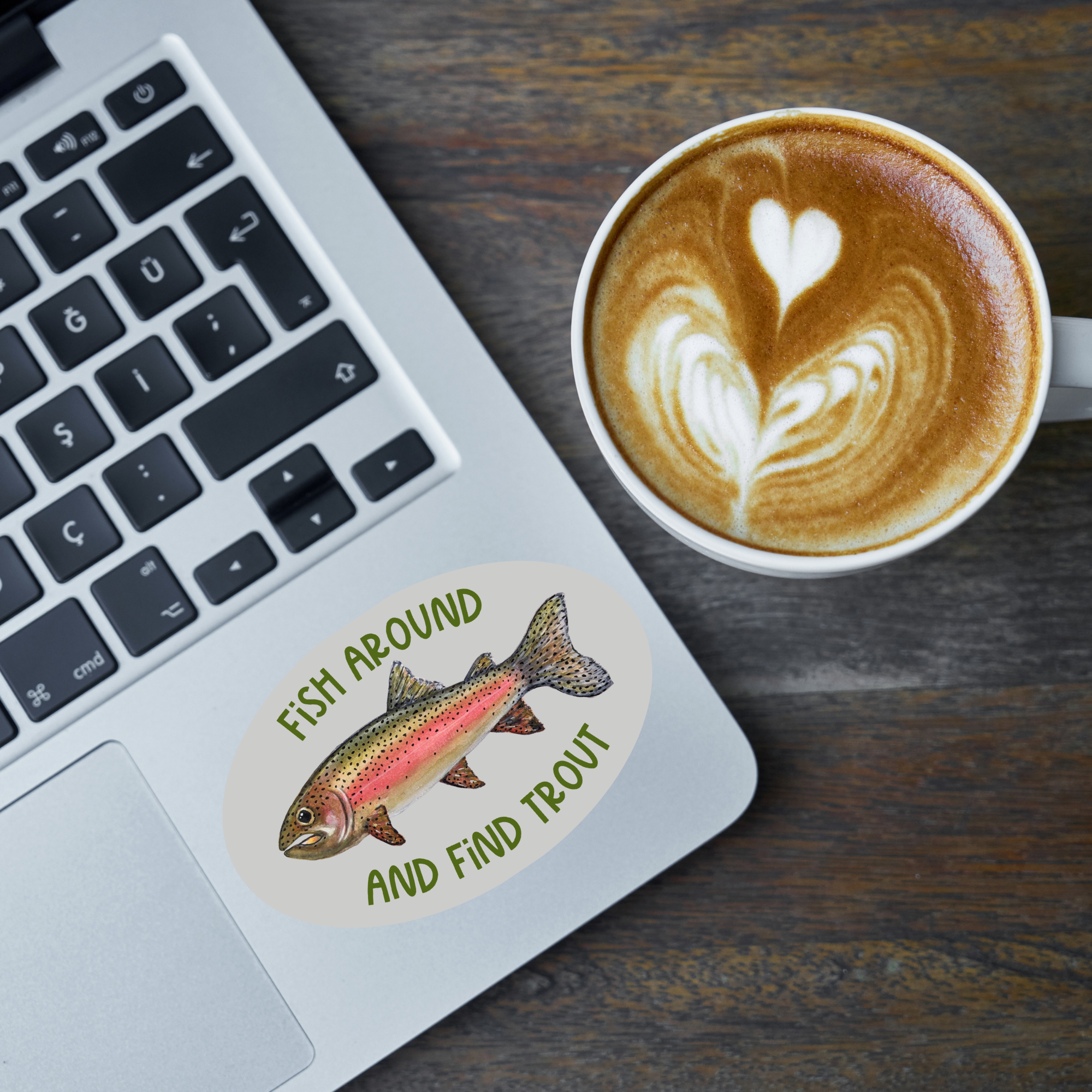 Oval-shaped sticker with a watercolor rainbow trout illustration and green text "Fish Around and Find Trout" on a grey background stuck on a laptop next to a coffee drink in a mug.