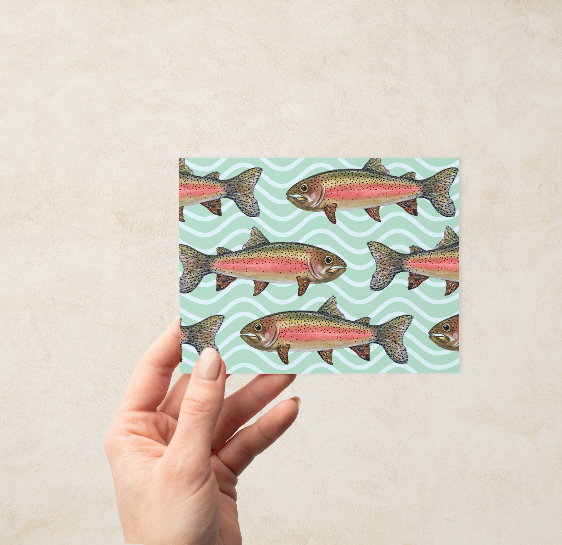 Hand holding a Greeting card with a school of watercolor rainbow trout illustrations on a mint green wavy pattern background.