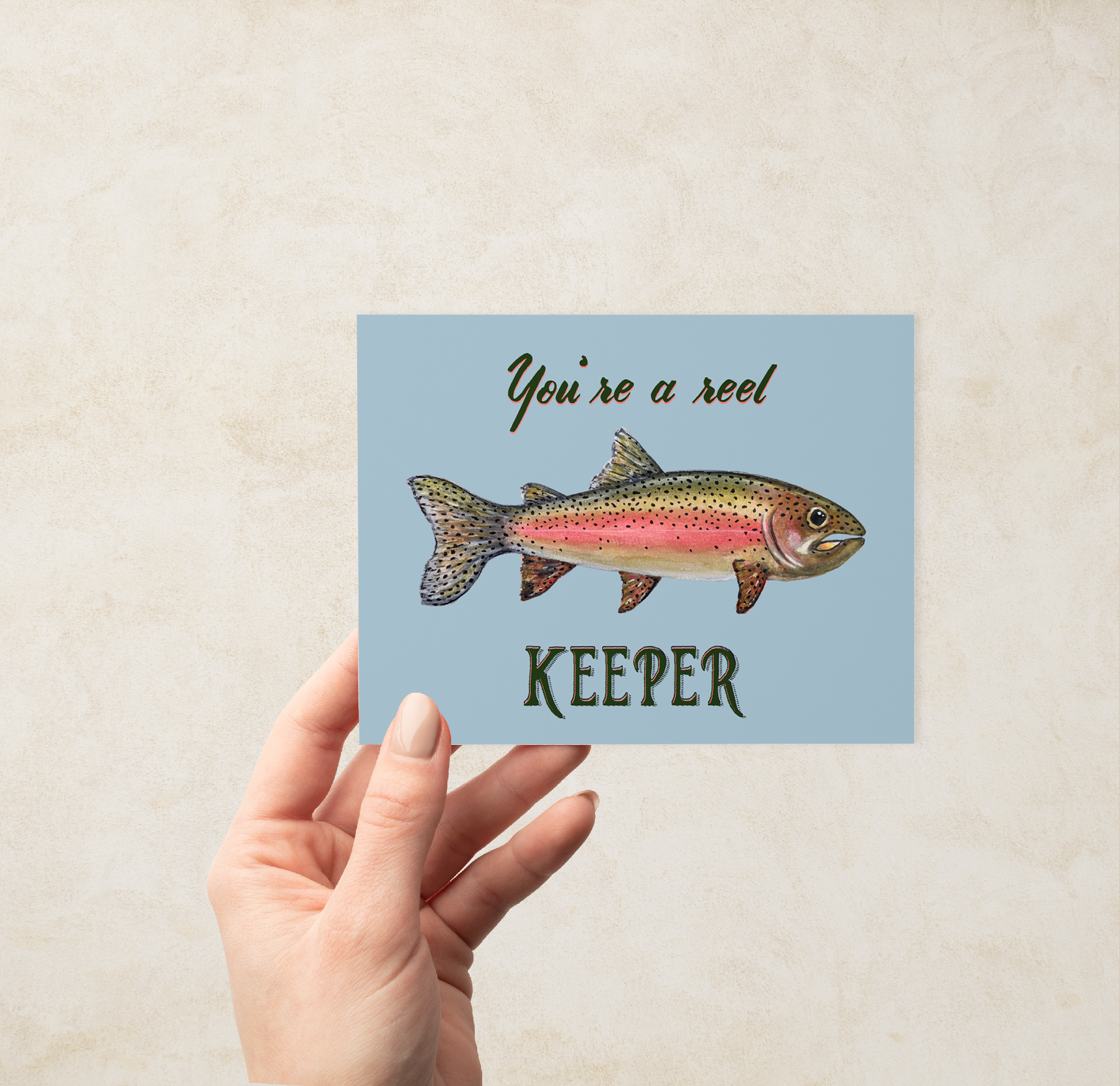 Hand holding a Greeting card with a watercolor rainbow trout illustration and text "You're a reel keeper" on a light blue background.