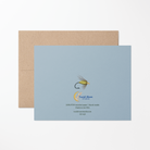 Back of the greeting card with a green fishing fly illustration, Lucid Moon Studio logo, website, and card information on a light blue background with a brown kraft envelope.