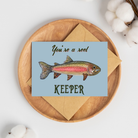 Greeting card with a watercolor rainbow trout illustration and text "You're a reel keeper" on a light blue background with a brown kraft envelope.