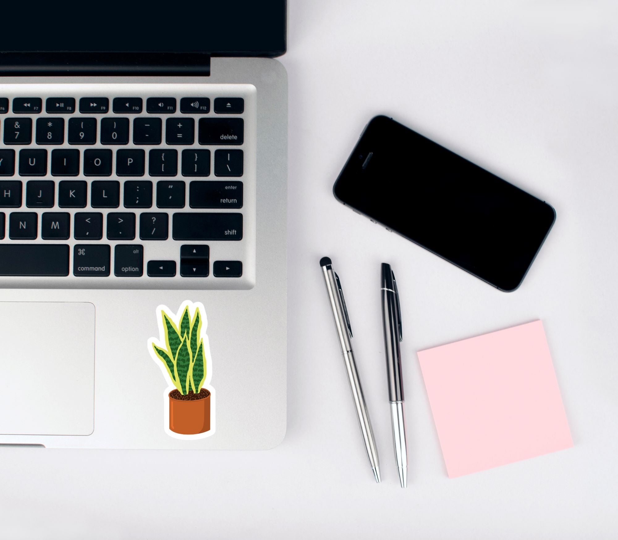 Vinyl matte sticker with a hand drawn illustration of a green and yellow snake plant in an orange terracotta pot against a white background on a laptop with a phone, pens, and notepad.