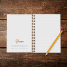 A photo of an opened wire spiral-bound notebook showing inside cover with Lucid Moon Studio logo and info about the notebook and lined pages on a wood background with a pencil.