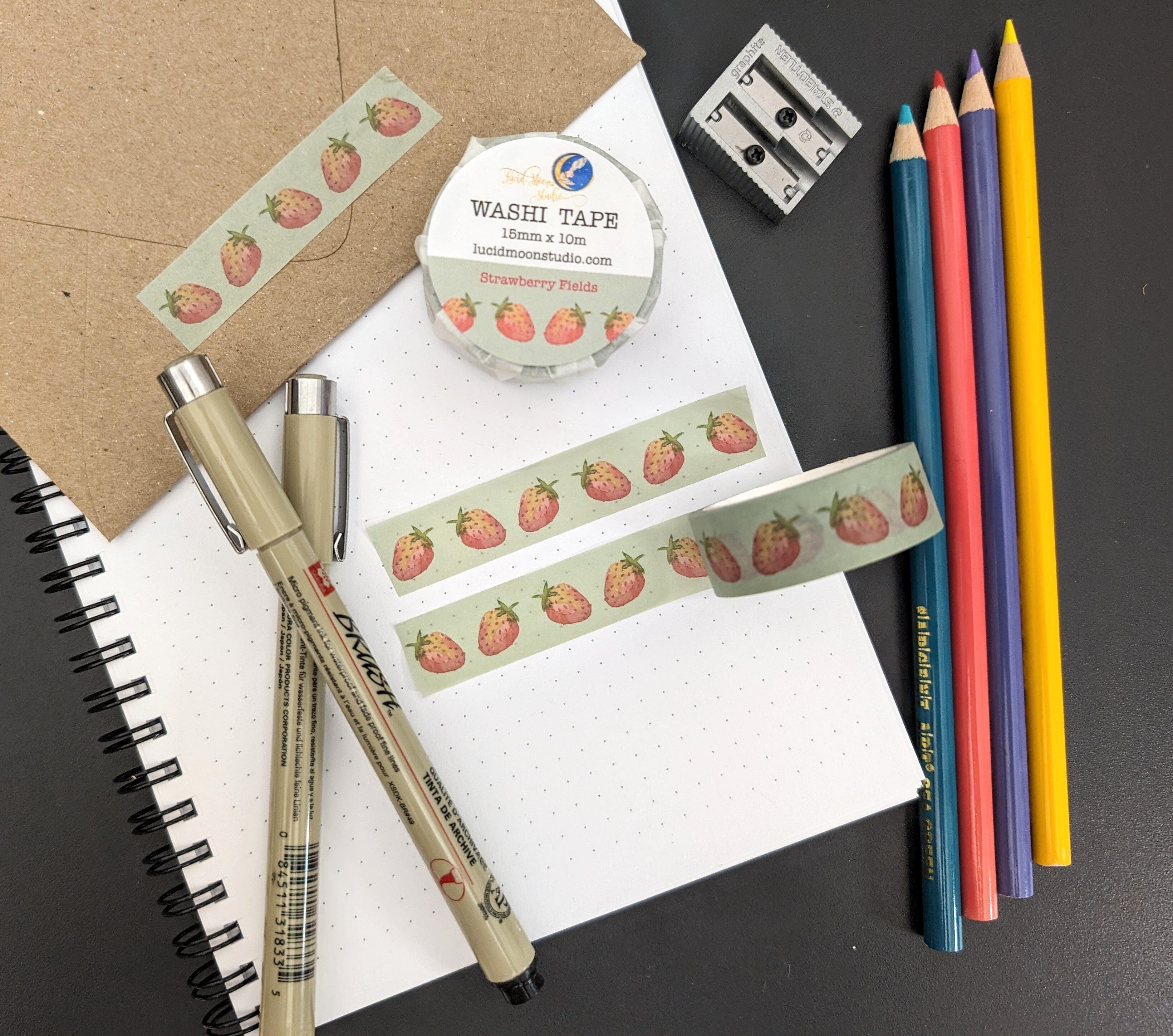 A roll of Strawberry Fields washi tape in a paper wrapper and 2 strips of the washi tape on a dot grid notebook and kraft envelope along with pens, colored pencils, and a pencil sharpener.