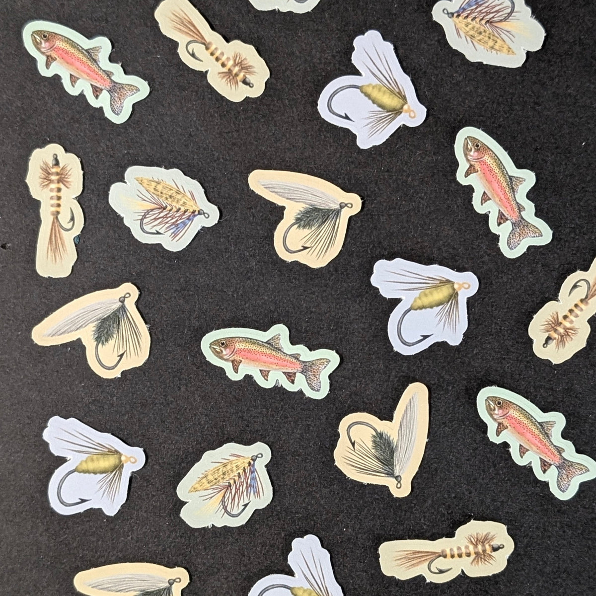 A collection of small illustrated fishing fly and rainbow trout stickers scattered on a dark textured background.