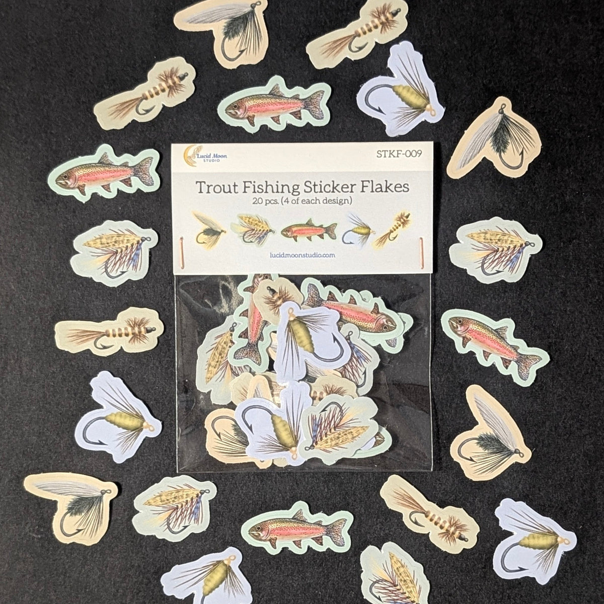 A pack of trout fishing sticker flakes arranged on a dark background, with the package label visible in the center and multiple fish, lure, and fly designs scattered around it.