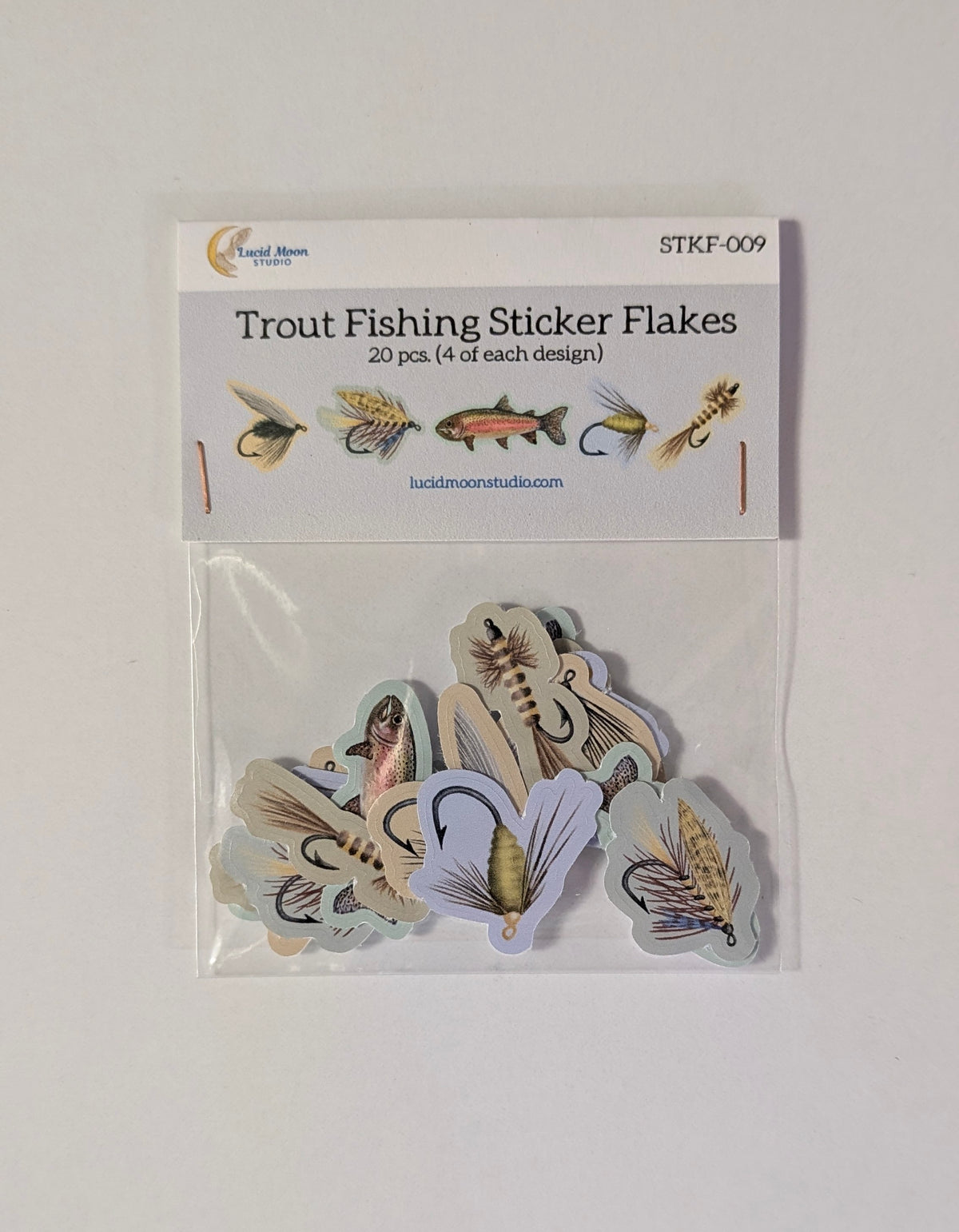 A clear plastic bag containing 20 trout fishing sticker flakes on a white backing card. The package is labeled 'Trout Fishing Sticker Flakes' with the brand 'Lucid Moon Studio' visible, and the flakes inside show various illustrated fish and fly lure designs in muted earthy tones.