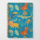 A photo of an 8.5" x 5.5" wire spiral-bound notebook featuring hand-illustrated cute jungle animals such as lions, tigers, toucans, iguanas, monkeys, and crocodiles on a bright blue background with green and brown leaves in a tossed pattern on the cover.